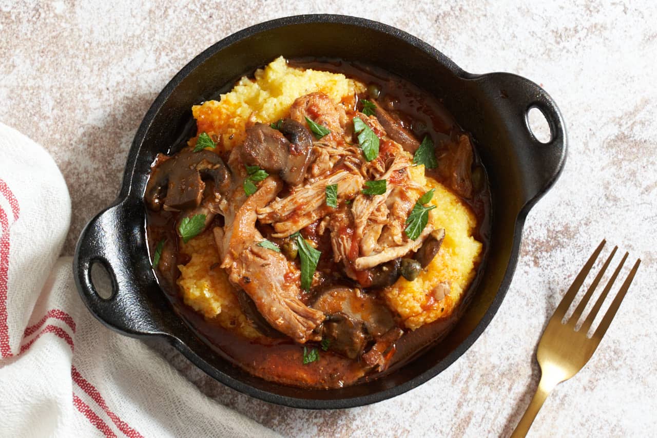 A cast iron serving dish of slow cooker chicken cacciatore over polenta. A gold forks is on the right, a red and white striped towel is on the left.