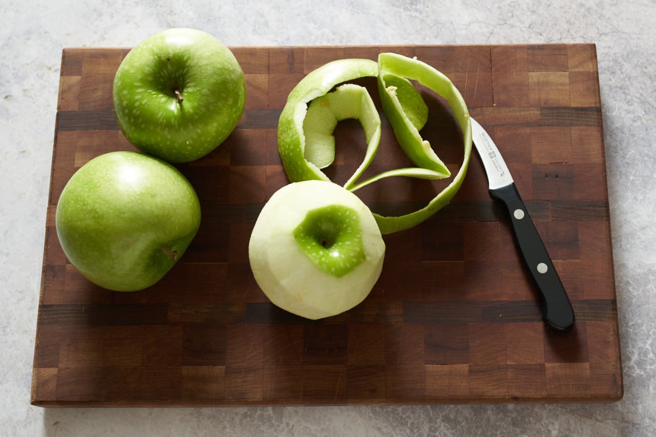 Three apples, one has been peeled, on a cutting board with a knife.