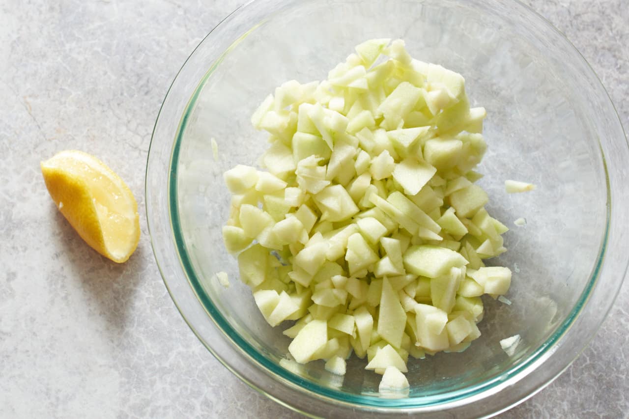 A bowl of chopped apple pieces next to a wedge of lemon that has been squeezed.