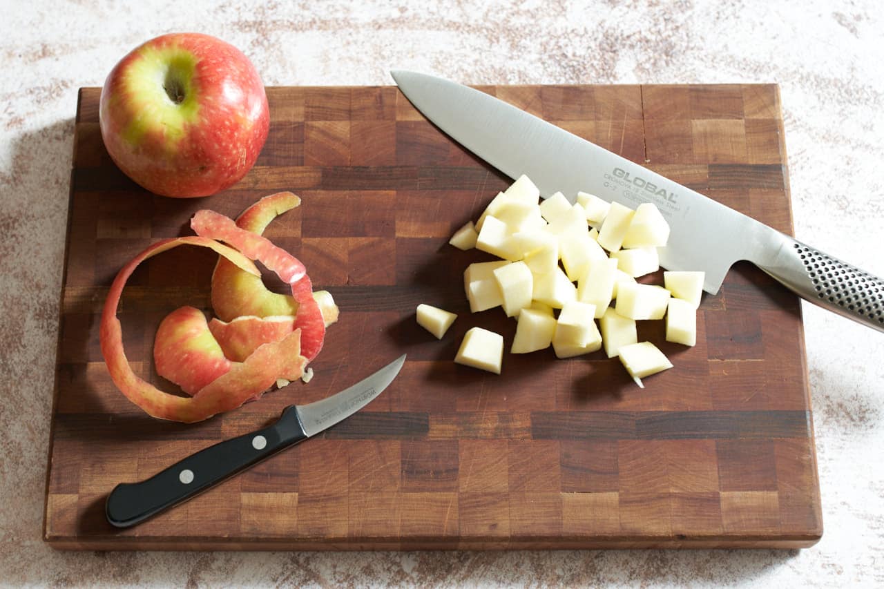 Chopped apple pieces on a cutting board with a knife, an apple and apple peels.