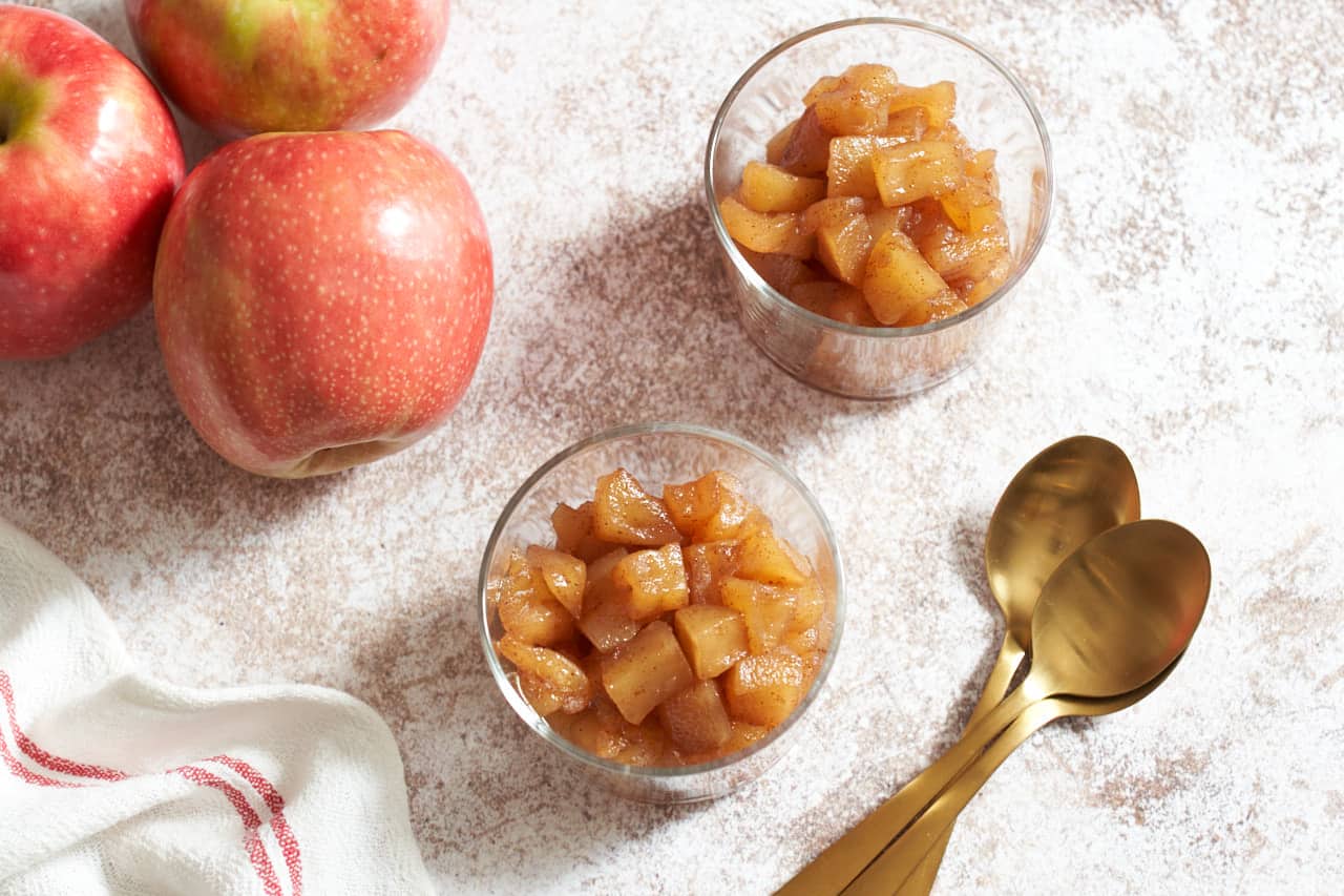 Two small glasses of apple compote. Three apples are in the upper left, a red and white striped towel is in the bottom left, and three gold spoons are on the right.