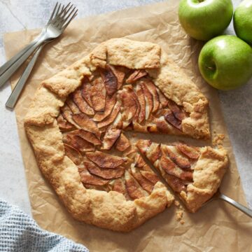 An apple galette with a slice cut into on a piece of parchment paper. A silver server is underneath the slice, three forks, a blue napkin and three apples surround the galette.