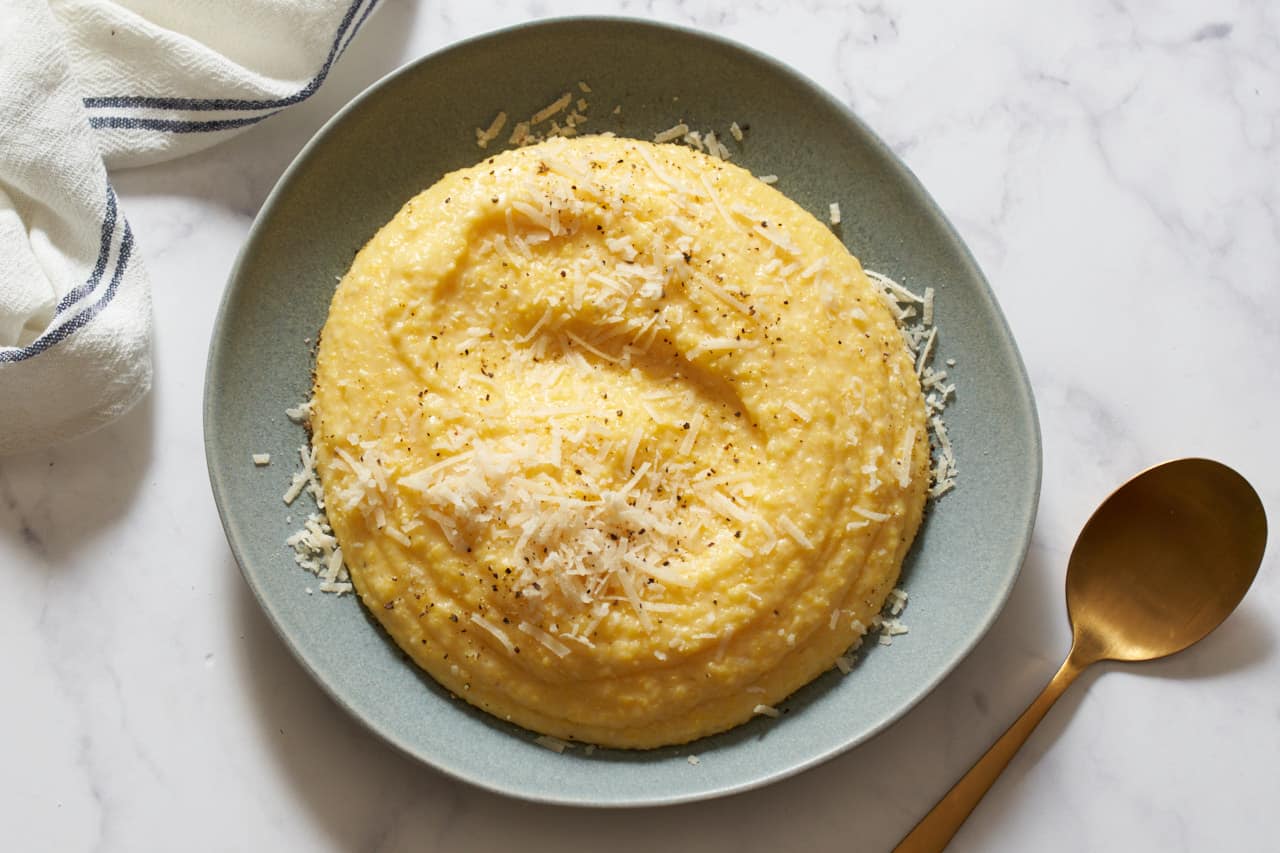 Creamy polenta topped with parmesan and black pepper on a gray plate. A gold spoon is on the right, a blue and white striped towel is on the left.