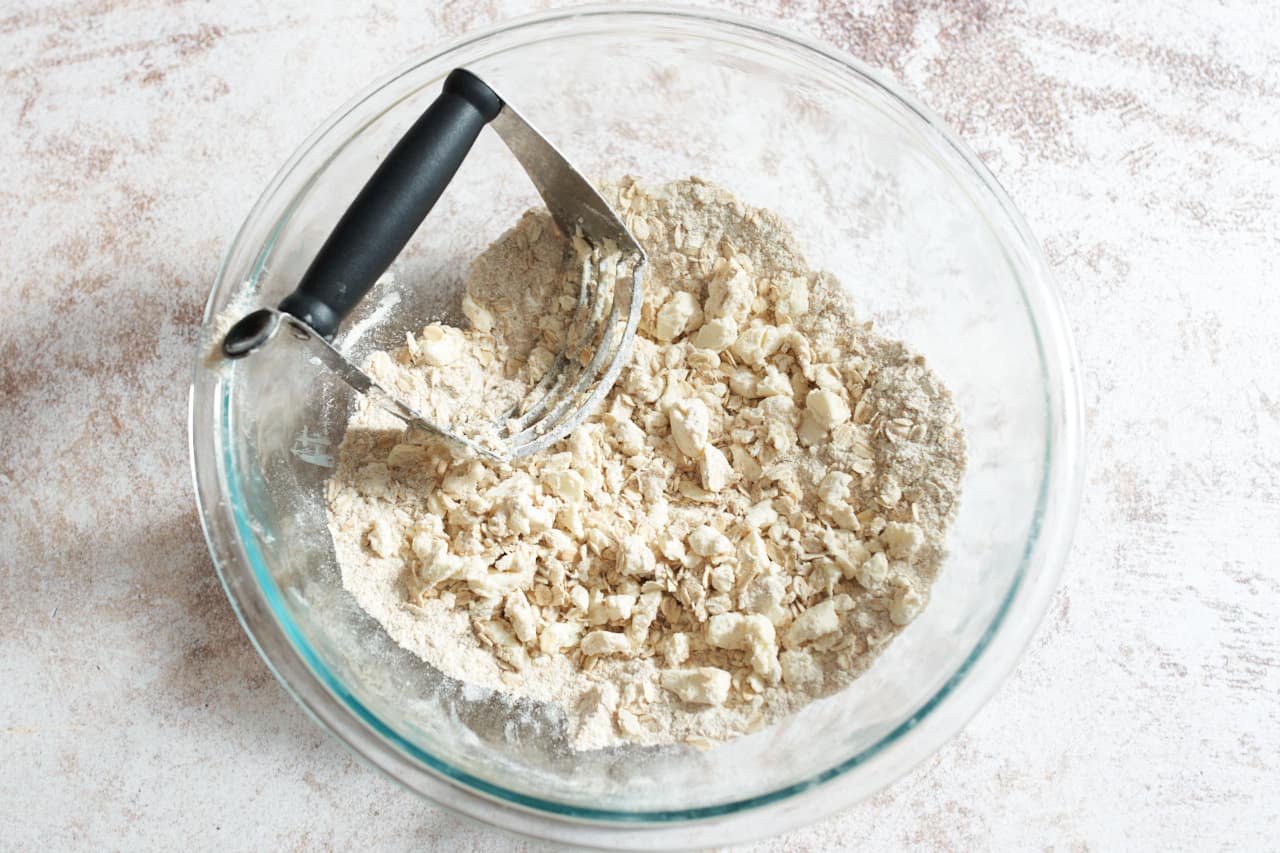 A pastry blender in a bowl shown working butter into dry ingredients for apple crisp topping.