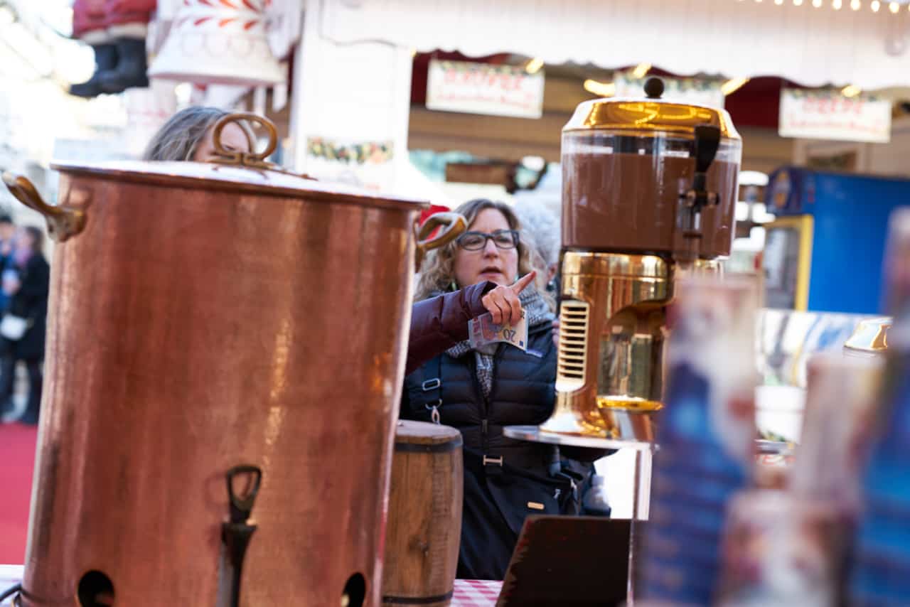 A woman at a booth selling mulled wine at a Christmas market in Paris. Large copper vats of wine are in the foreground.