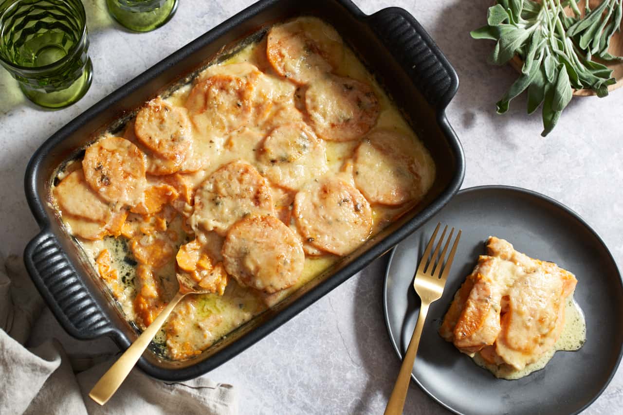 A sweet potato gratin with a gold spoon in it, next to a portion of the gratin on a black plate with a gold fork. A bowl of sage is in the upper right corner, two green glasses are in the upper left.