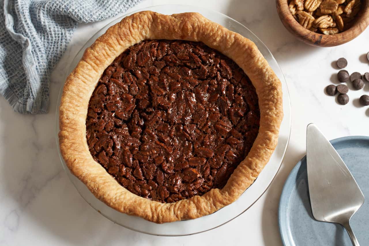 A chocolate pecan pie in a glass pie dish. A blue napkin is in the upper left, pecans, chocolate chips, and a blue plate with a pie server are on the right.