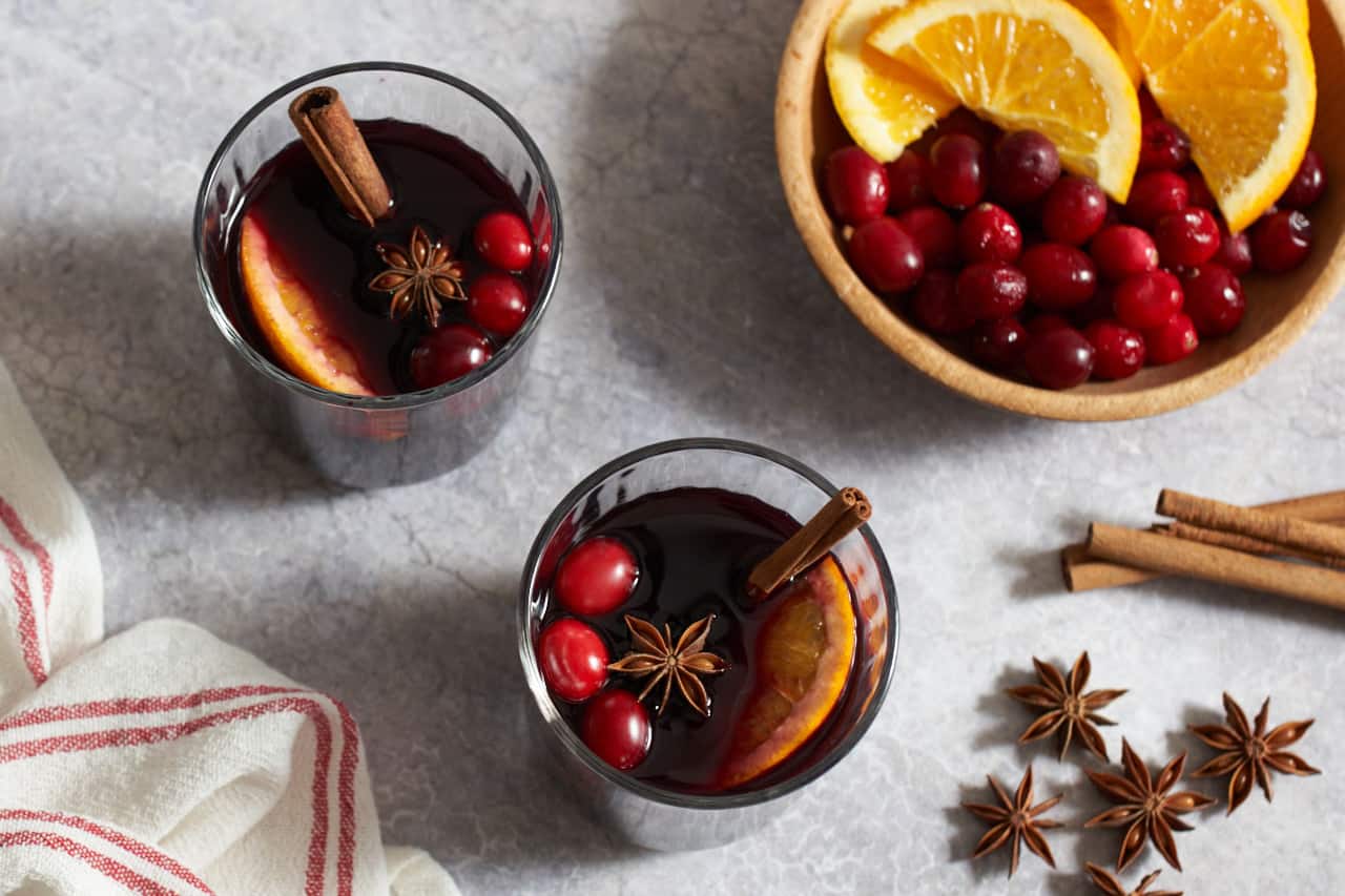 Two glasses of mulled wine garnished with star anise, orange slices, fresh cranberries and cinnamon sticks. A red and white striped towel is on the bottom left, a bowl of cranberries and oranges, cinnamon sticks and some star anise pods are on the right.