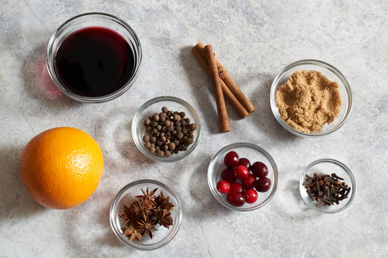 An orange, three cinnamon sticks, and small glass bowls with red wine, fresh cranberries, star anise, whole allspice, whole cloves, and brown sugar.