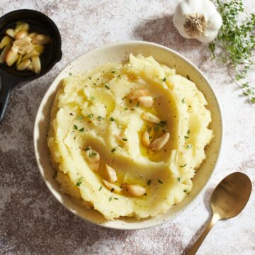 A bowl of mashed potatoes topped with roasted garlic cloves and fresh thyme. A tiny cast iron skillet filled with roasted garlic cloves and olive oil is in the upper left, a garlic bulb and a bunch of thyme are in the upper right, a gold spoon is on the lower right.