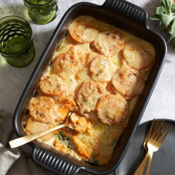 A sweet potato gratin with a gold spoon in it, next to a black plate with gold forks. A bowl of sage is in the upper right corner, two green glasses are in the upper left.