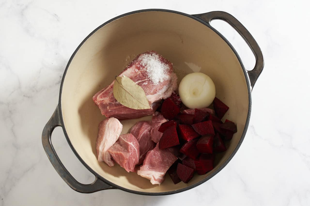 A Dutch oven with chopped pork, an onion, a bay leaf and diced beets.