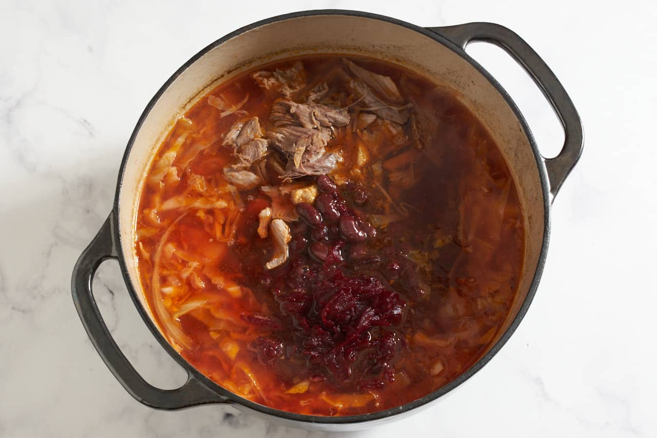 Cooked beets, pork, and kidney beans being added to a pot of borscht soup.