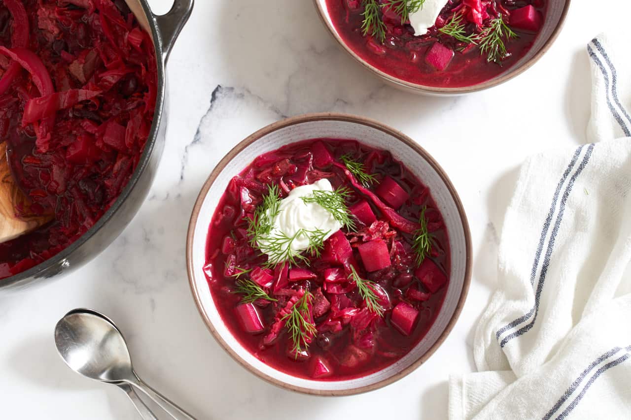 Two bowls of borscht soup topped with sour cream and fresh dill. A pot of borscht is on the left with two spoons and a blue and white striped towel is on the right.