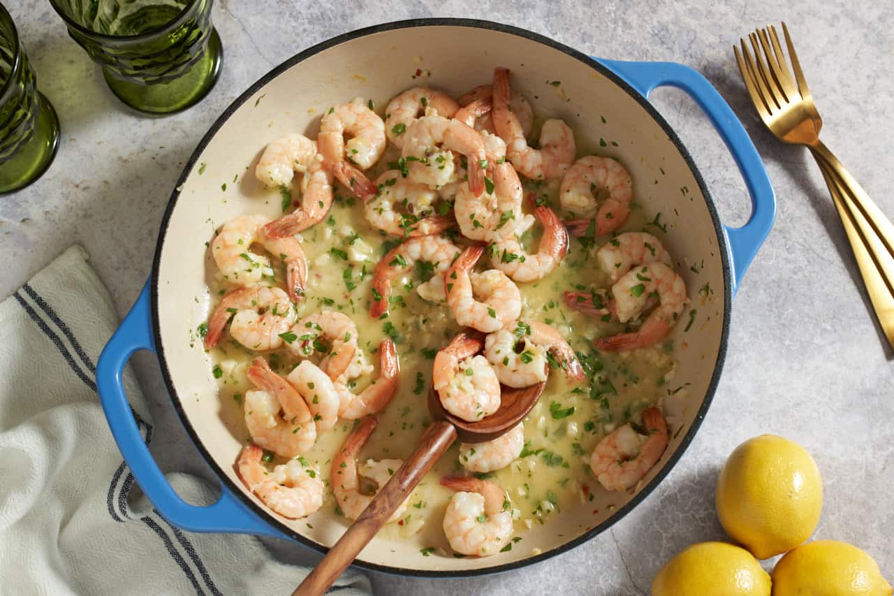 Shrimp scampi in a pan with blue handles with a wooden spoon in it. The pan is surrouned by two green glasses, three gold forks, three lemons, and a blue and white striped napkin.