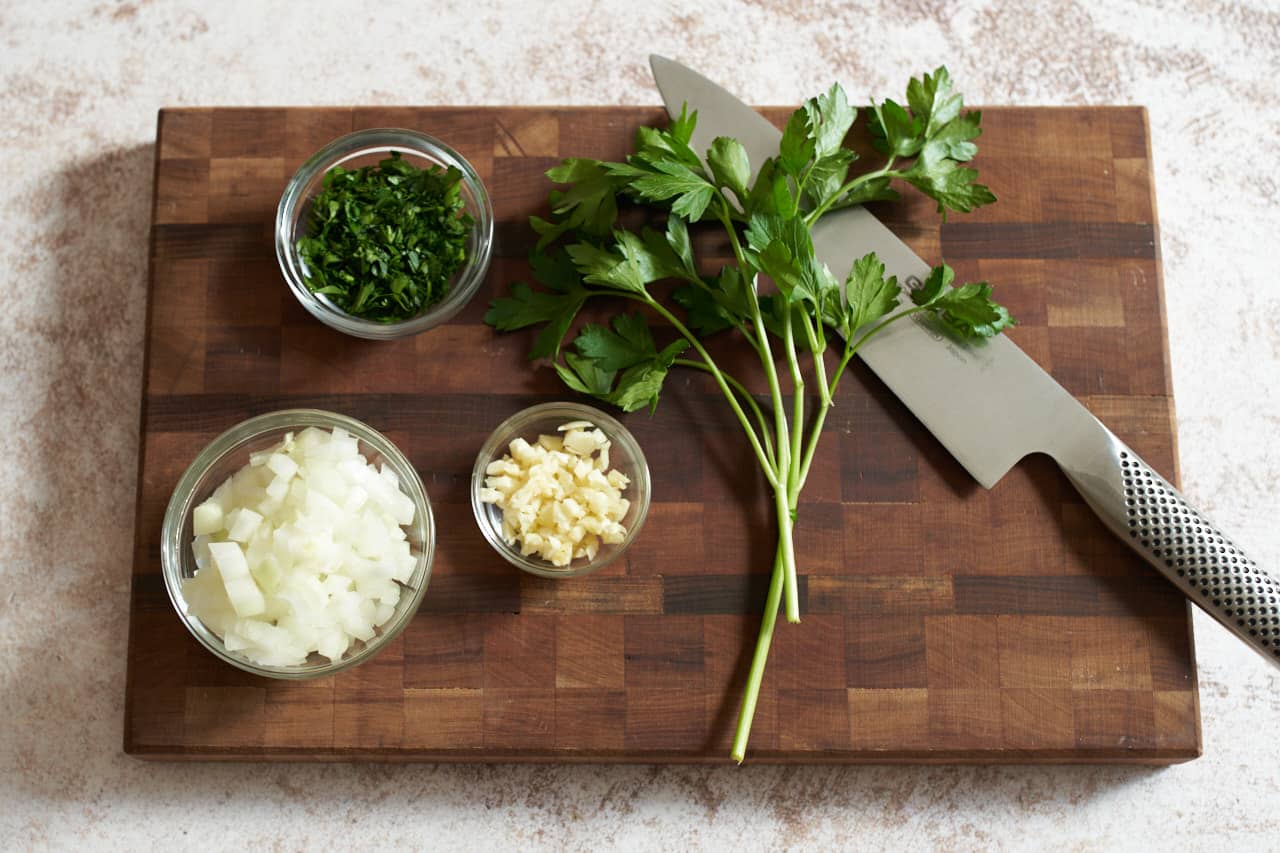 A knife on a cutting board next to three small bowls of minced onions, parsley, and garlic.