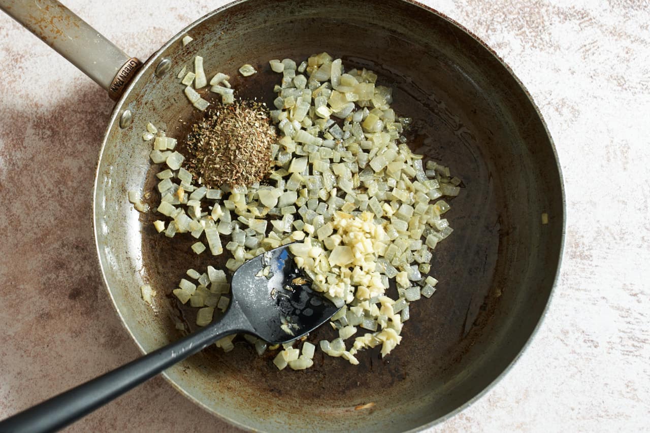 A skillet with softened onions, dried Greek oregano, and minced garlic.