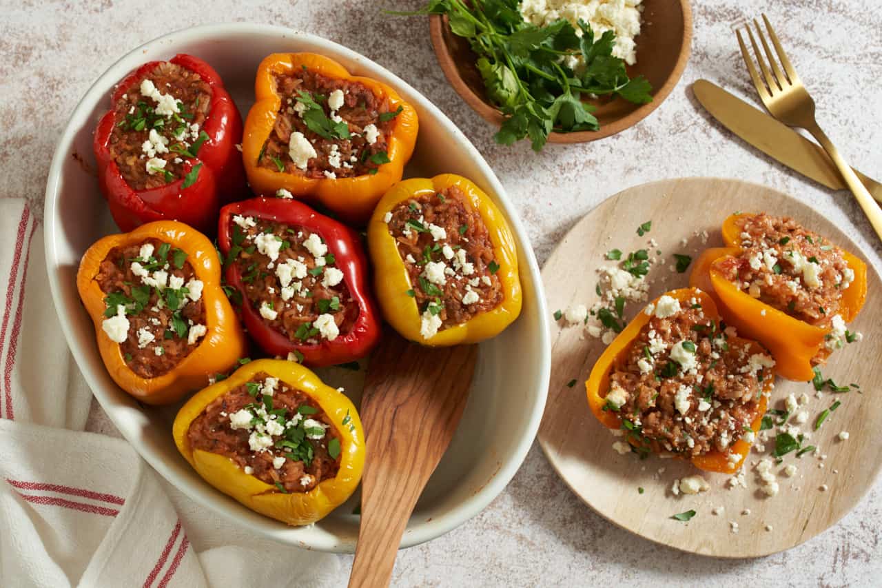 Greek stuffed peppers topped with feta and parsley in an oval baking dish with a wooden spatula in it. A plate with a sliced pepper is on the right, a gold knife and fork and a small bowl with parsley and feta are above it.