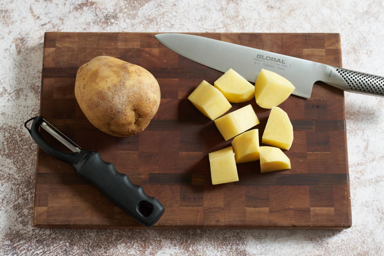 A knife and a vegetable peeler on a wooden cutting board with chopped potatoes.