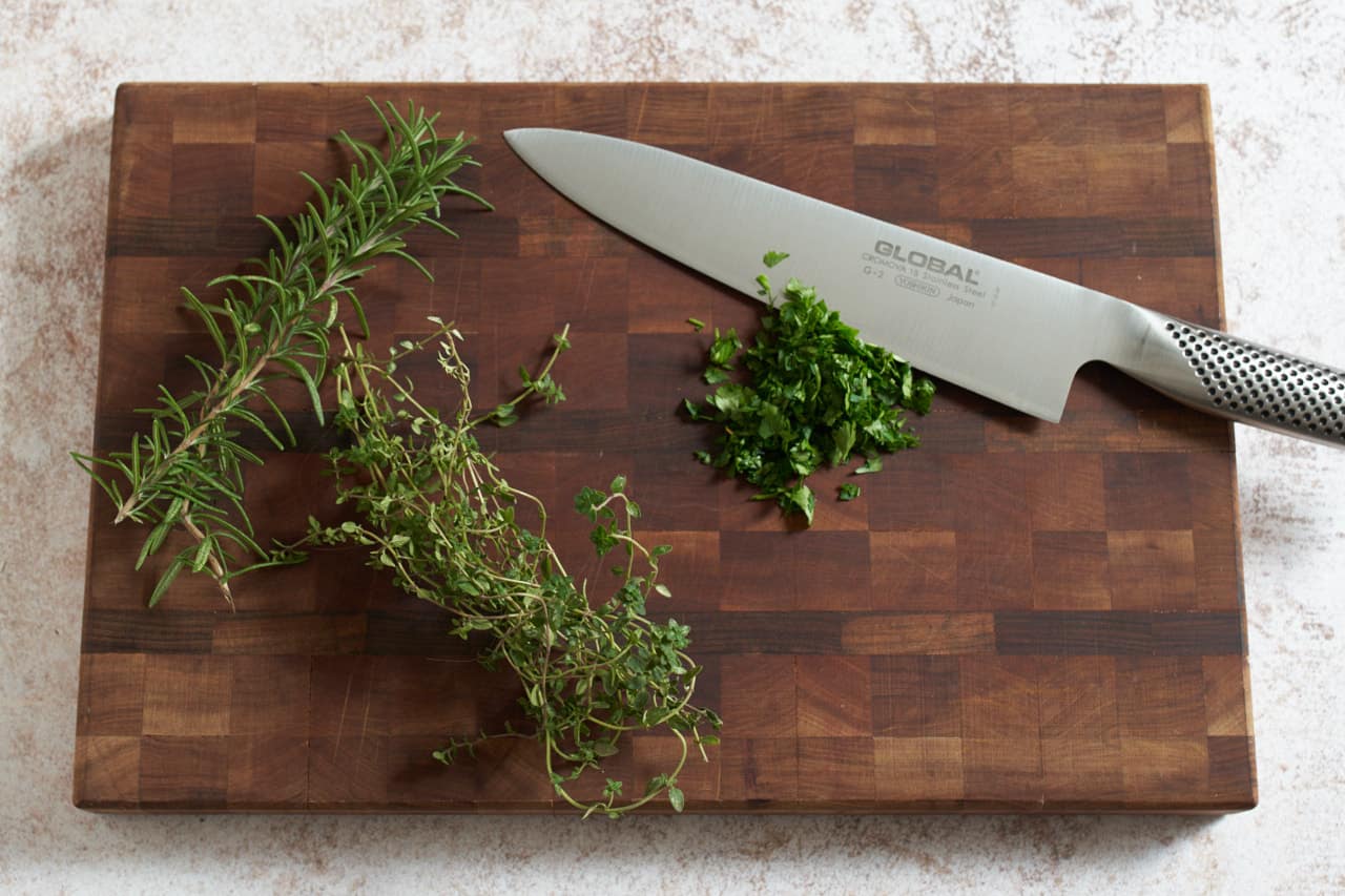A knife on a cutting board with fresh herbs being chopped.