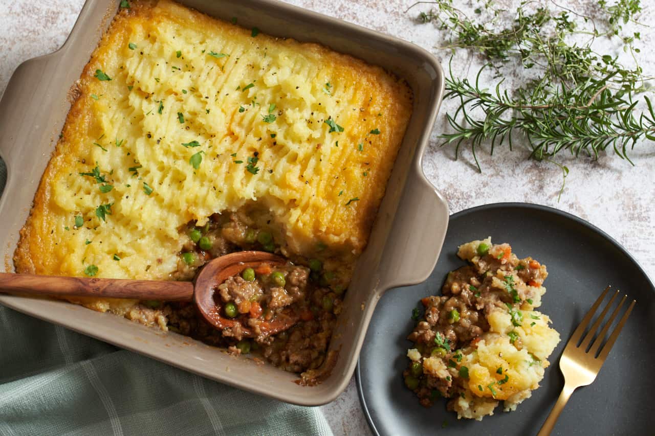 A square pan of shepherd's pie with a wooden spoon in next to a black plate with a portion of shepherd's pie and a gold fork. A green towel is on the left and fresh thyme and rosemary are in the upper right.