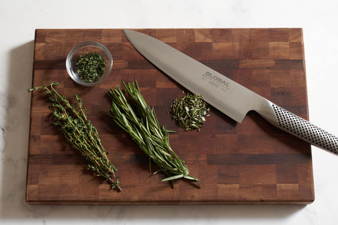 A knife on a cutting board with fresh rosemary and thyme.