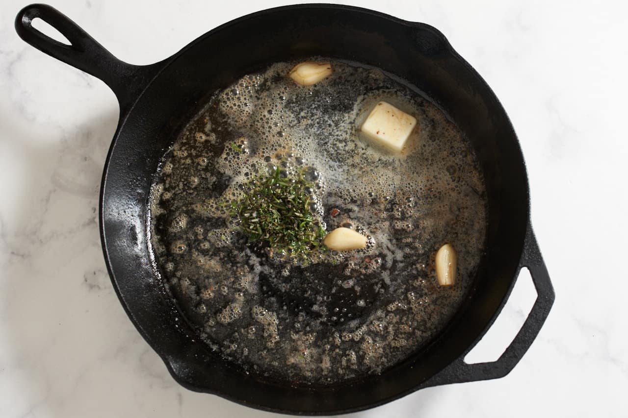 Foaming butter, chopped fresh herbs and garlic cloves in a cast iron skillet.