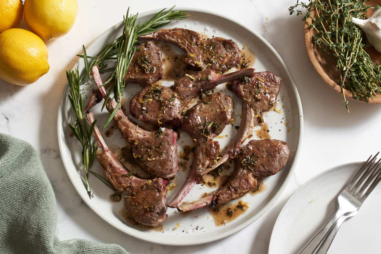 Pan seared lamb chops on a white plate with fresh sprigs of rosemary. Fresh lemons, a green towel, a bowl with herbs and garlic, and a plate with two forks surround it.