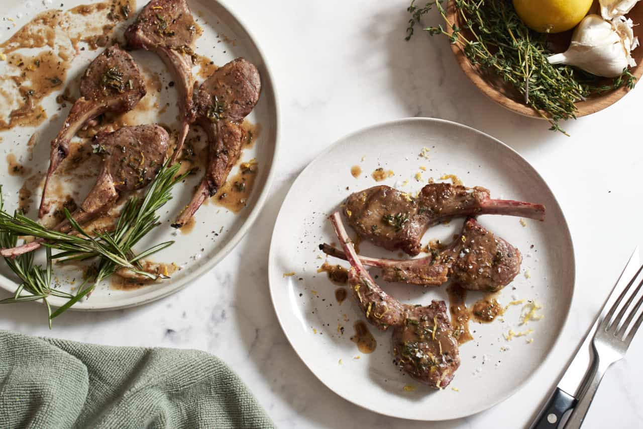 Two plates of pan seared lamb chops. A green towel is on the left, a bowl of herbs and garlic and a fork and knife are on the right.