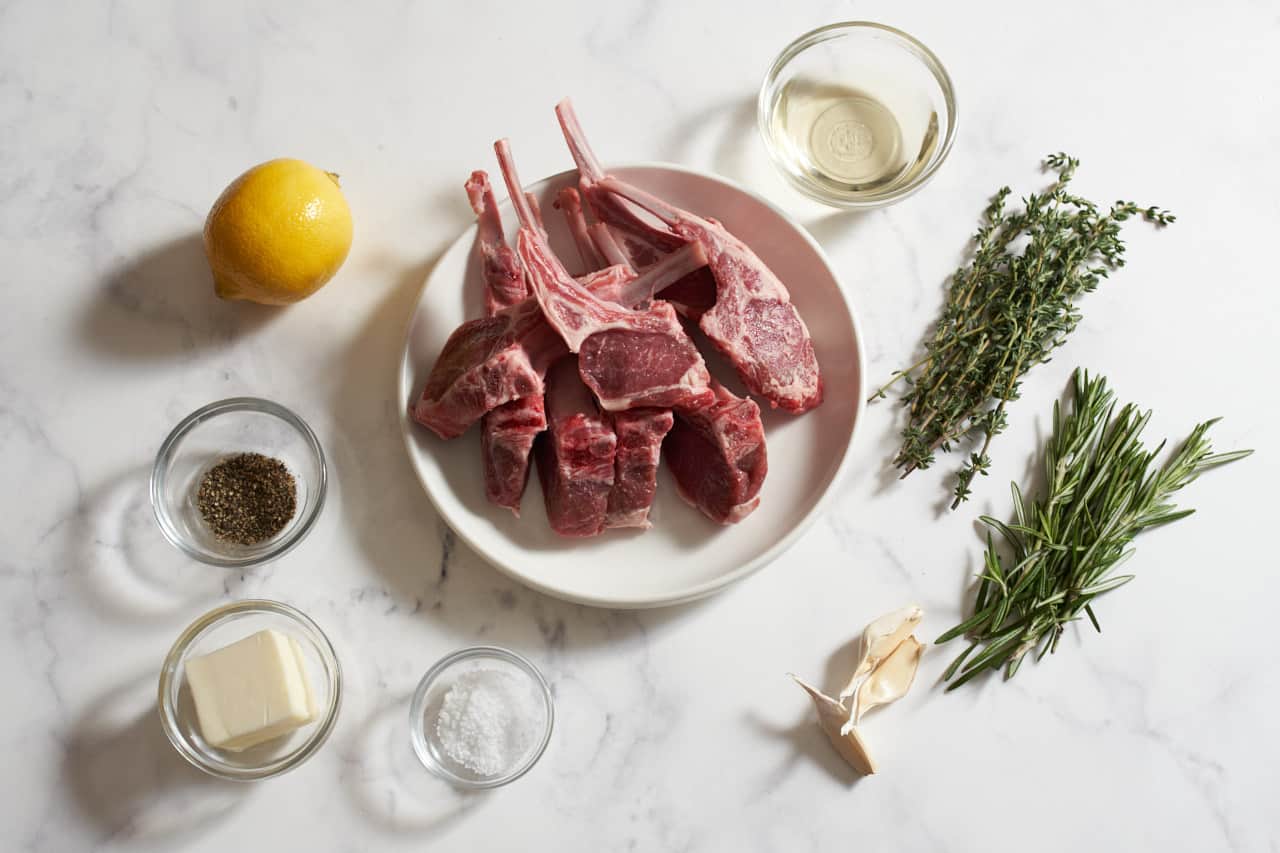 A plate of lamb chops surrounded by fresh thyme, fresh rosemary, a lemon, garlic cloves, and small bowls of salt, pepper, butter, and white wine.