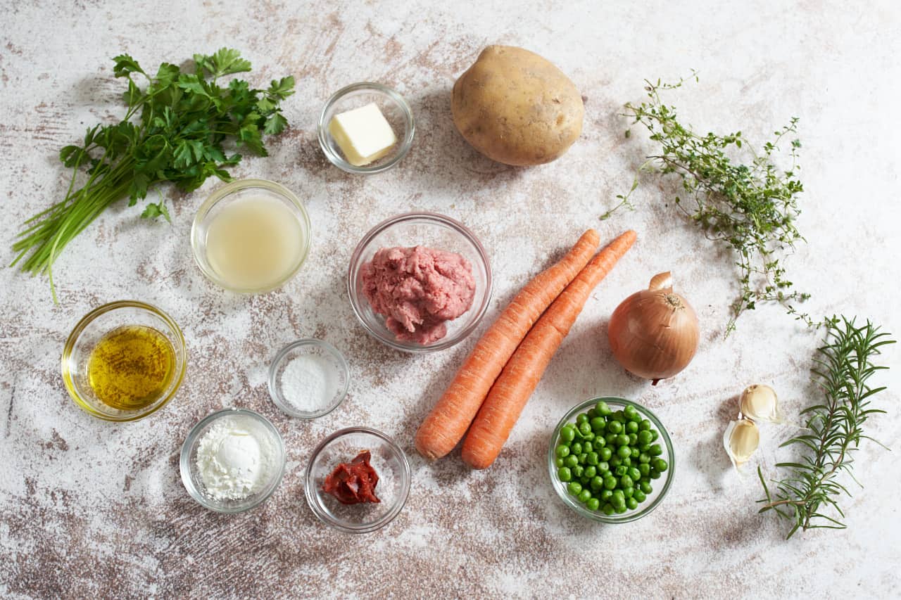 Two carrots, an onion, a potato, garlic cloves, fresh rosemary, parsley and thyme, and small bowls of ground lamb, salt, olive oil, corn starch, butter, peas and broth.