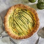 An asparagus gruyere quiche with a blue and white striped towel on the left, a pie server and two green glasses are on the right.