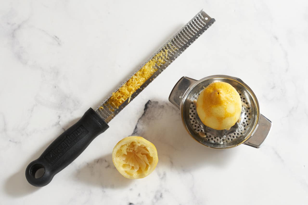 A microplane grater with lemon zest on it next to a juicer and half a lemon.