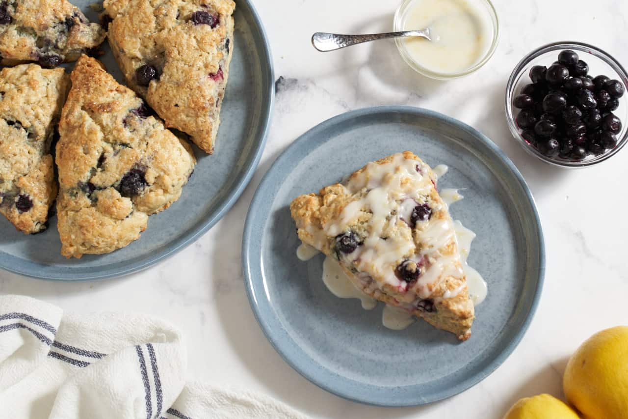 A glazed lemon blueberry scone on a blue plate. A plate of more scones is in the upper left, two small bowls with glaze and blueberries are on the right.