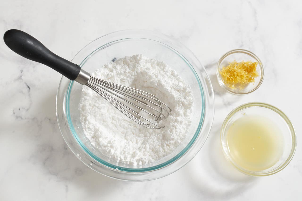 A bowl of powdered sugar with a whisk in it next to small bowls of lemon juice and zest.