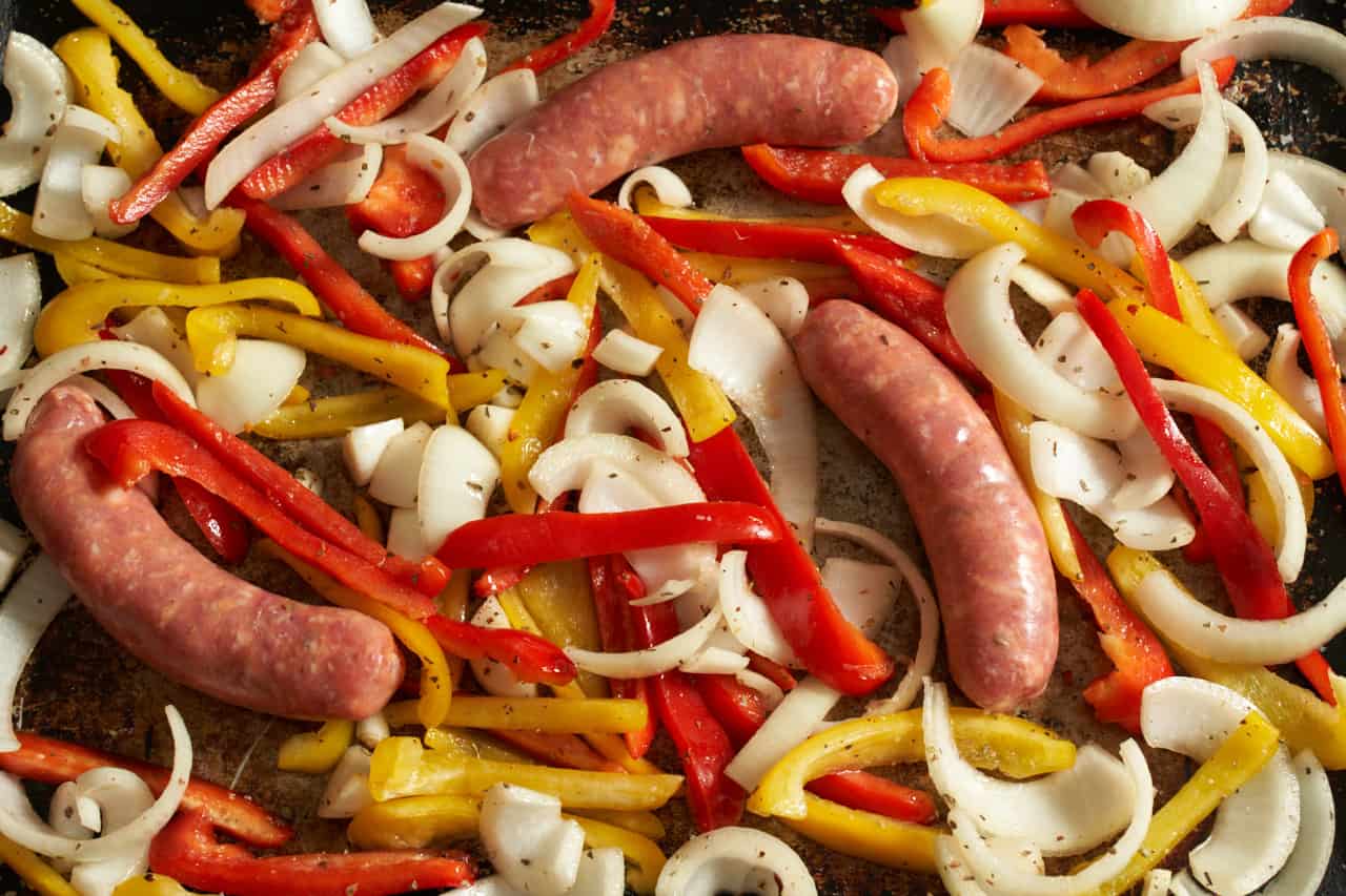 Sliced red and yellow peppers and onions with Italian sausage on a sheet pan.