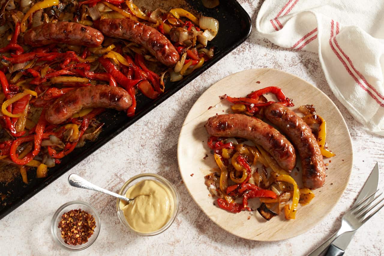 Oven baked sausage and peppers and onions on a sheet pan and a plate. A red and white striped towel is in the upper right. Small bowls of condiments and some utensils are on either side of the plate.