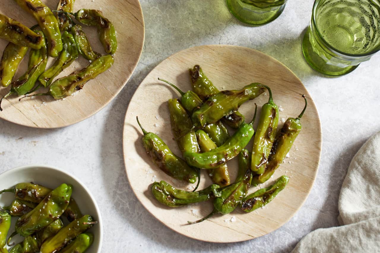 Two plates and a bowl of blistered shishito peppers, two green glasses and a tan towel are on the right.