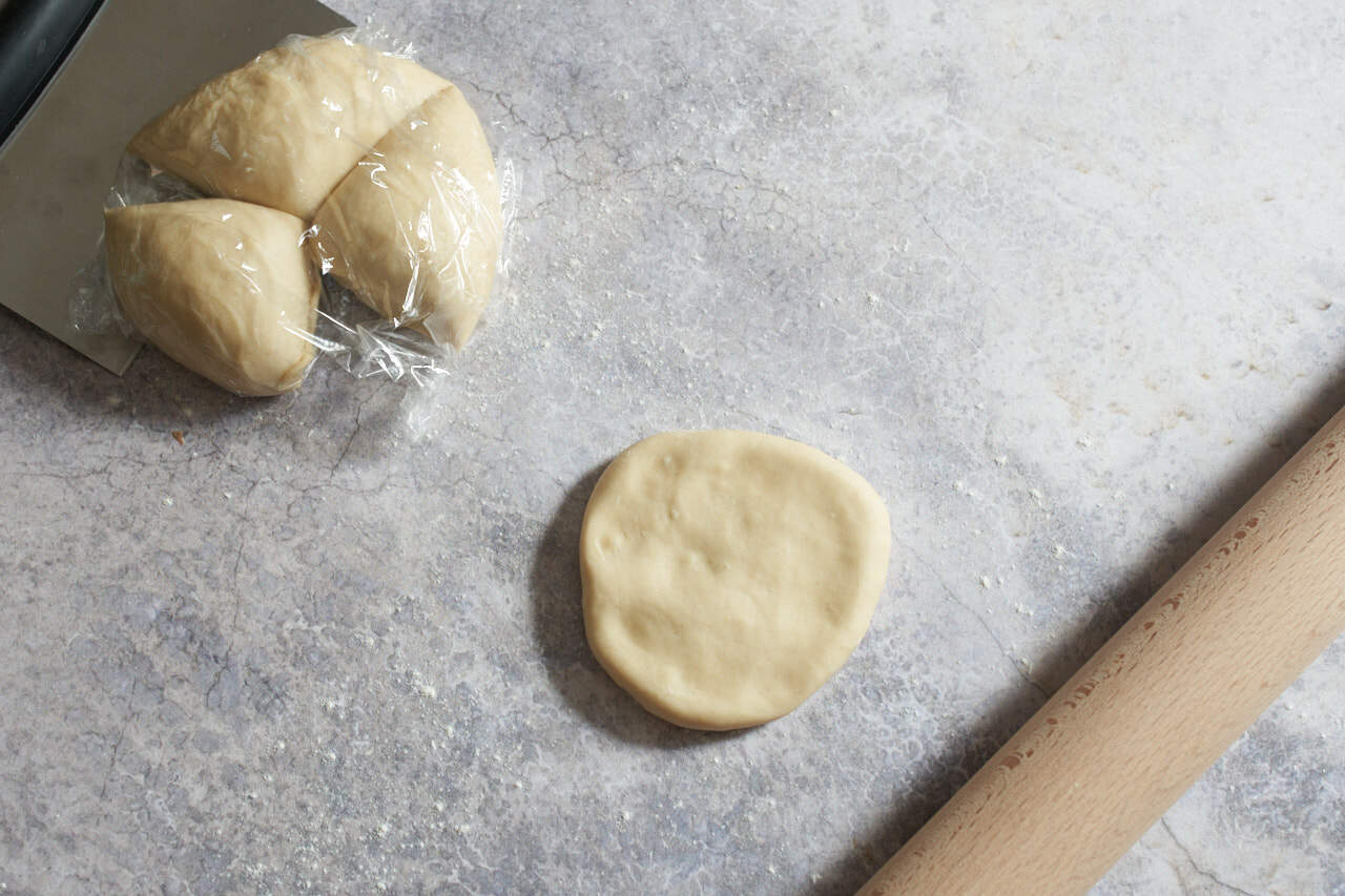 Varenyky dough cut into three pieces and covered in plastic is in the upper left. A disc of dough is in the center, and a rolling pin is on the right.