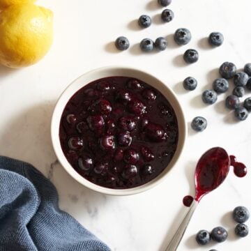 A bowl of blueberry compote, a spoon that has been dipped in the compote is on the right, fresh blueberries, a lemon and a blue towel surround the bowl.