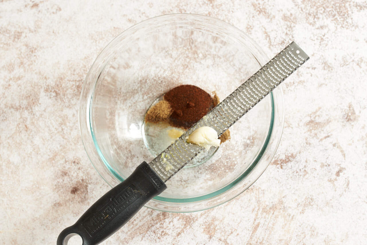 A microplane grater with a garlic clove over a bowl of taco seasoning.