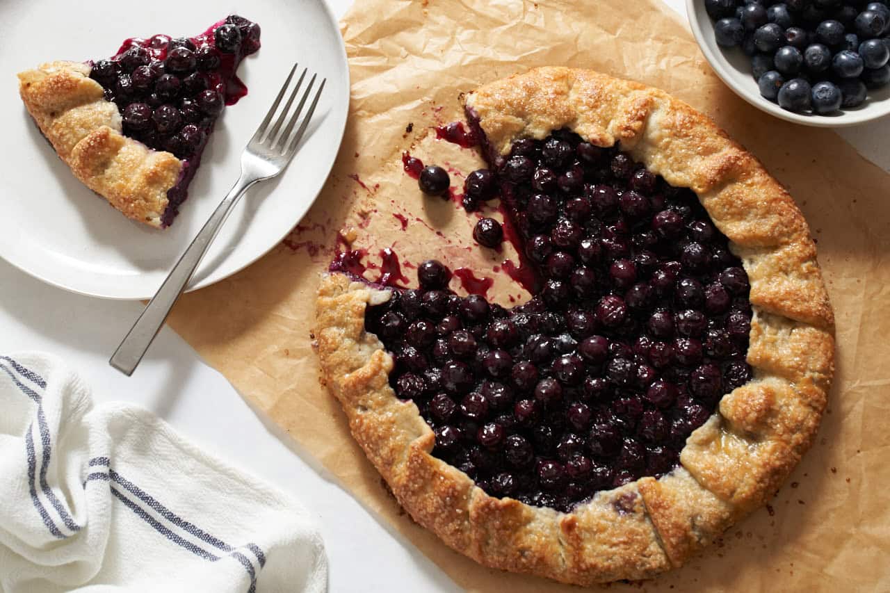 A blueberry galette with a slice cut out of it on parchment paper, a slice of the galette is on a plate to the left, a bowl of blueberries is on the right.