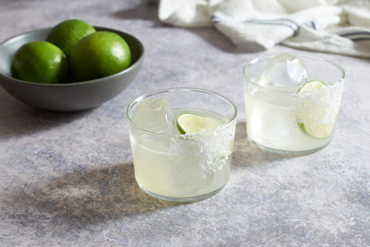 Two mezcal margaritas with salt, a bowl of limes and a blue and white striped towel are in the background.