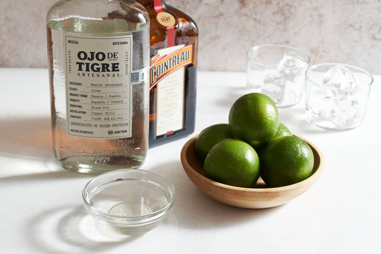 Bottles of mezcal and Cointreau next to a bowl of limes, a bowl of simple syrup, and two glasses filled with ice.