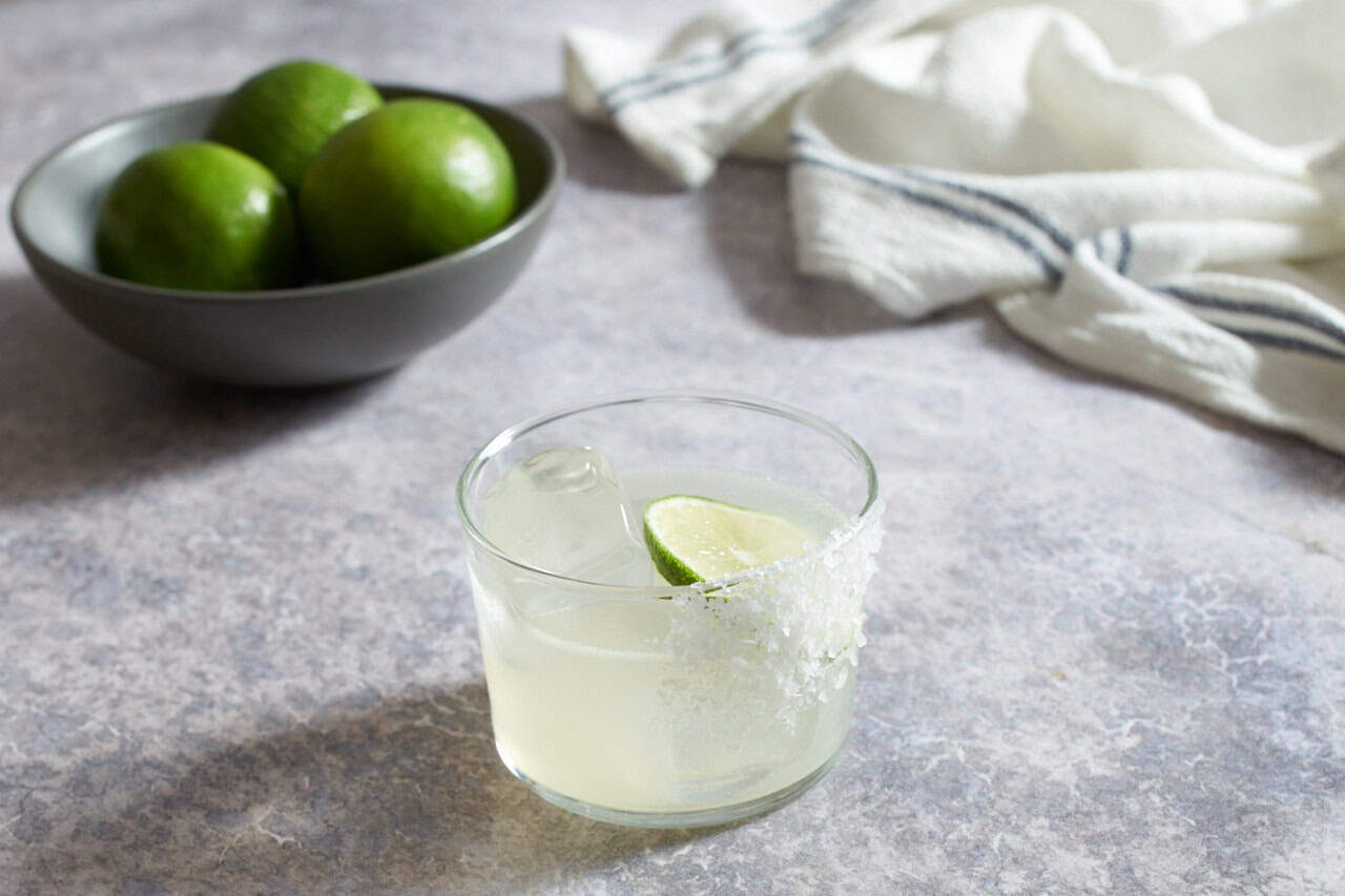 A mezcal margarita with salt, a bowl of limes and a blue and white striped towel are in the background.