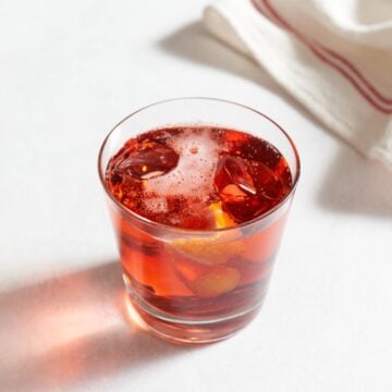 A negroni sbagliato drink in a glass with ice and a slice of orange zest, with a red and white striped towel in the background.