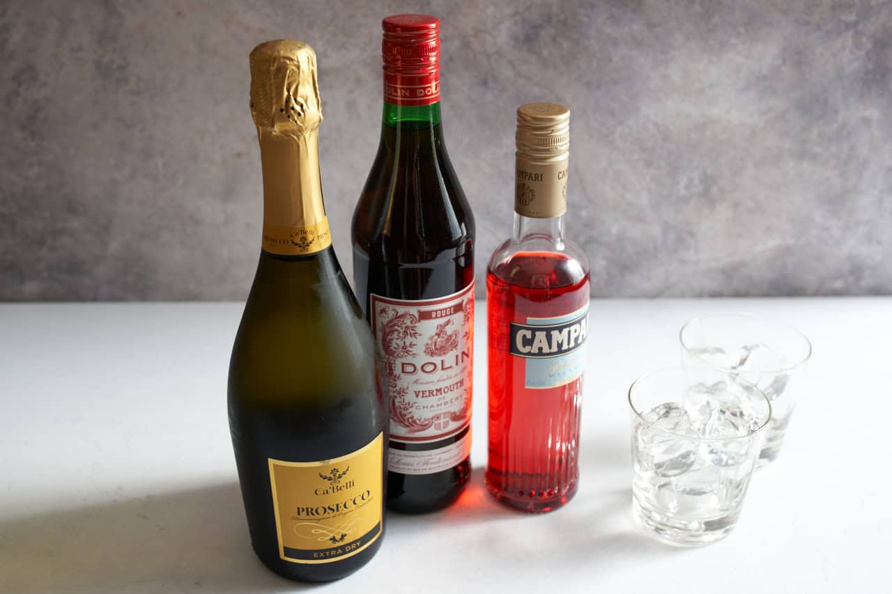 A bottle of prosecco, a bottle of sweet vermouth, and a bottle of Campari alongside two cocktail glasses full of ice.