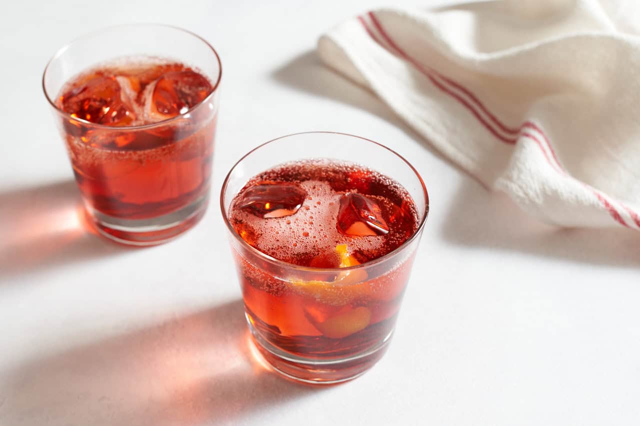 A negroni sbagliato drink in a glass with ice and a slice of orange zest, with a red and white striped towel, and another drink in the background.