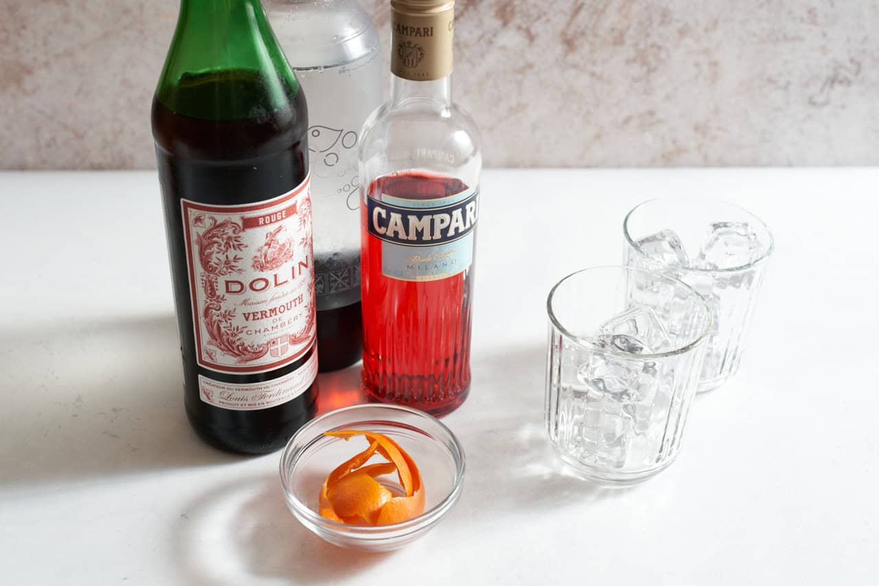 Bottles of Campari, sweet vermouth, and club soda next to a bowl of orange peels and two glasses full of ice.