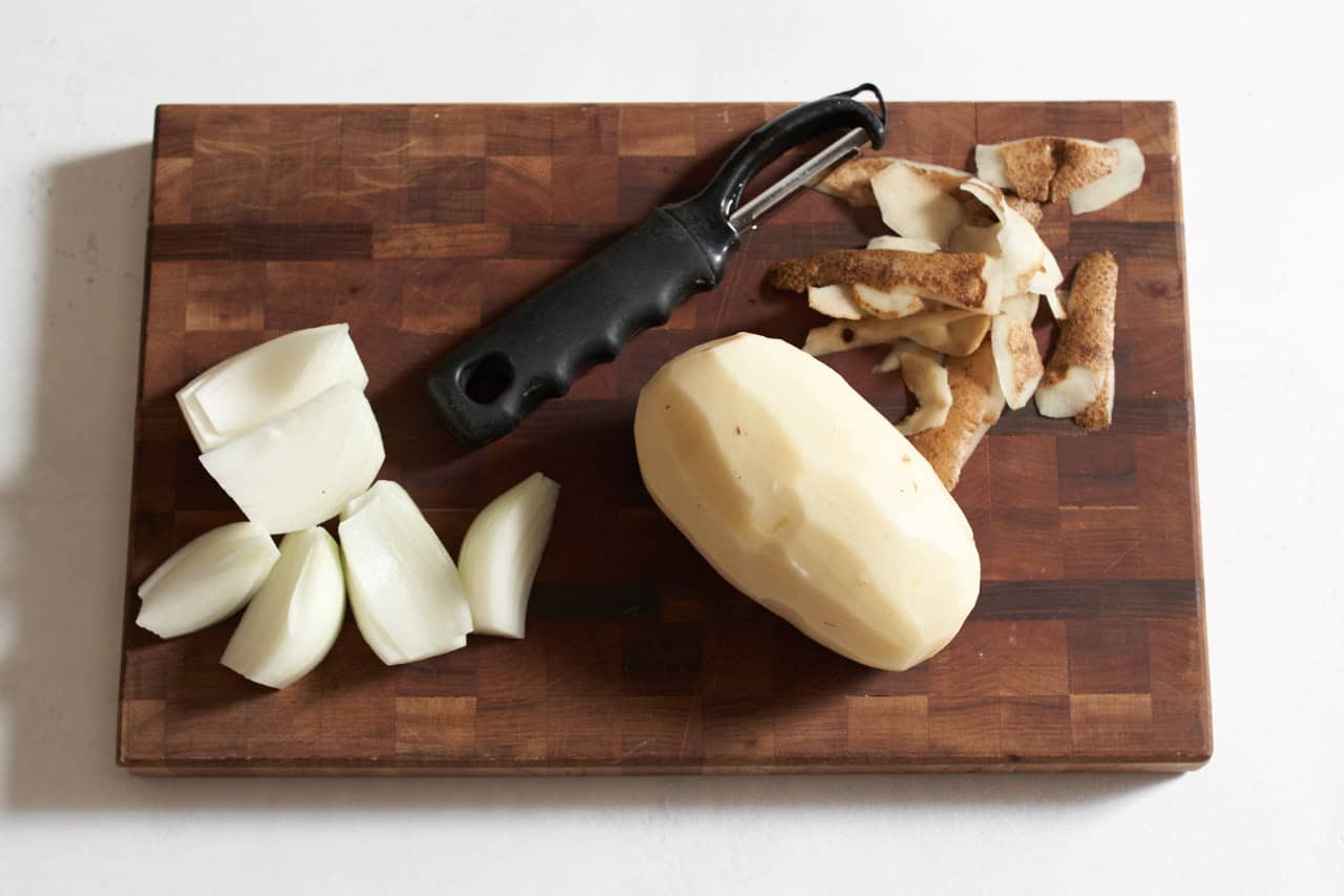 A vegetable peeler on a wooden cutting board with a peeled potato and a quartered onion.